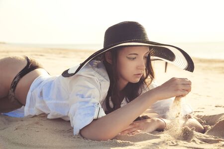 beautiful young woman in hat and blouse at sand sea beach under light rays lying plays with sandの写真素材