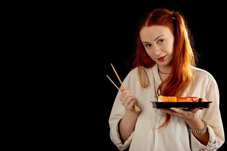 happy red hair young woman with sushi on black background with copy space looking at cameraの写真素材