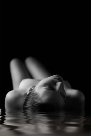 beautiful young aroused nude woman with closed eyes in dark lying on water, monochromeの写真素材