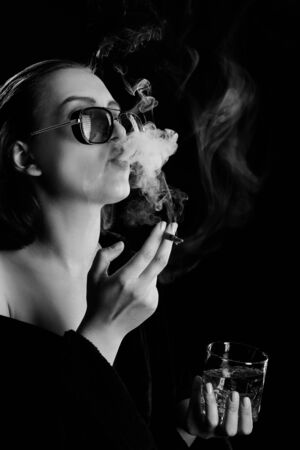 serious young woman in bathrobe and sunglasses, bare shoulder, whiskey in glass and cigarette, blow up smoke, monochromeの写真素材