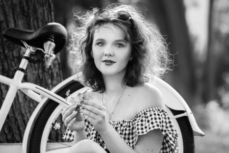beautiful pinup girl with bare shoulders dress and curly red hair sitting with bike in sunny park in sun light, looking at camera, monochromeの写真素材