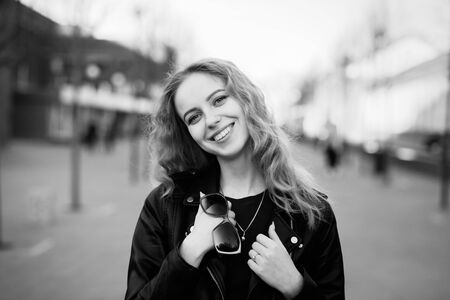 attractive curly blond girl in leather jacket standing on city street looking at camera, smiling,...
