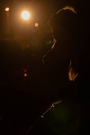 young woman standing near wall, embraced herself with closed eyes, dark background with back light...