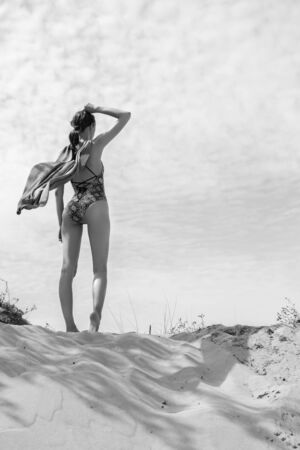 young woman in swimwear and cloak stand on sand dune under wind looking at horizon, rear view, monochromeの写真素材