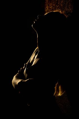 young woman standing near wall, embraced herself with closed eyes, dark background with back light, outdoors, profile viewの写真素材
