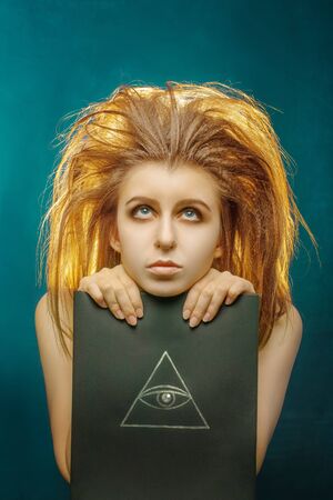 sad young woman with long fluffy red hair and big black book with mental Mason sign on green background looking upの写真素材