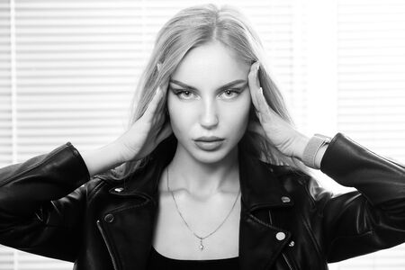 sensual blond girl in leather jacket with hands on head, looking at camera, jalousie background,...