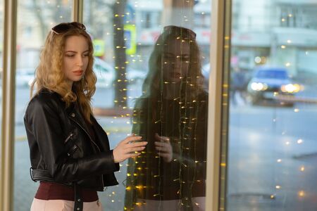 serious cute blond woman in leather coat stand on street near yellow garland looking at camera