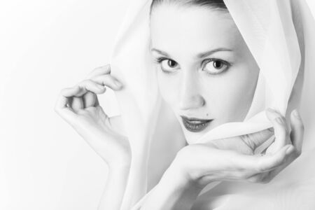 beautiful young woman with eastern makeup cover her head with white veil on white background, looking at camera, smiling, monochromeの写真素材