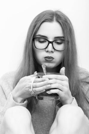 young woman in eyeglasses sitting on white background blows mug of hot tea, monochromeの写真素材