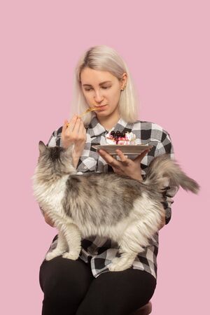 happy pretty blond girl with sweet dessert on pink background looking at her cat smilingの写真素材