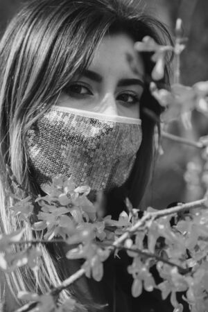 serious young woman with medical protective mask at spring flowers looks at camera, monochromeの写真素材