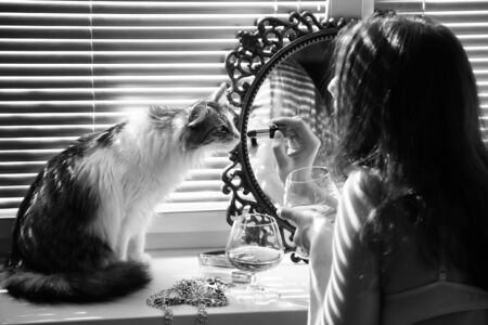 beautiful young woman with her cat applying cosmetics near mirror in sun light, monochromeの写真素材