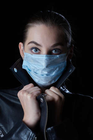 young serious woman in medical mask and leather coat at dark looks at cameraの写真素材
