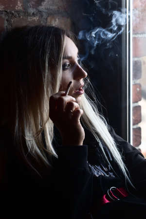 sad young luxury blond woman sitting near window, smoking, looking away