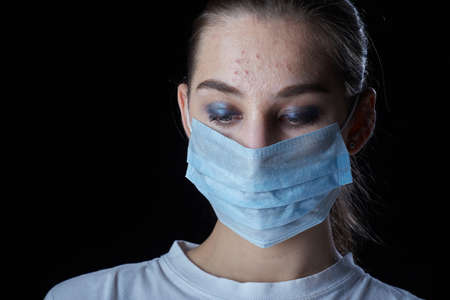 young sad woman with acne in medical mask on black background looks downの写真素材