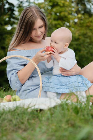 young happy mom with her baby on hands sitting at grass in park, smiling, baby eats apple