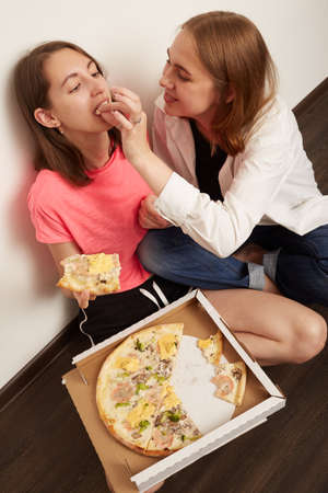 Two happy lesbian girls eating pizza indoors while sitting on the floorの写真素材