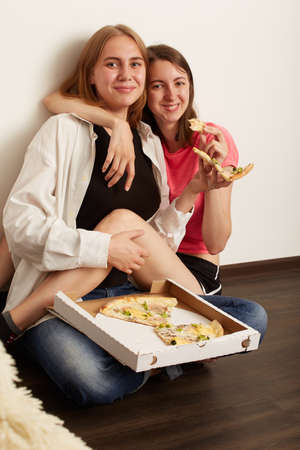 Two happy lesbian girls eating pizza indoors while sitting on the floorの写真素材