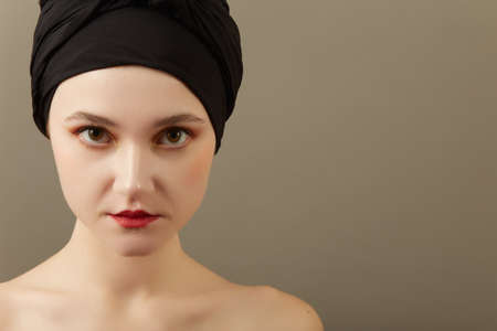 sensual young woman with bare shoulders and luxury professional makeup in headscarf on gray background with copy space looking at cameraの写真素材