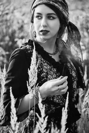 young gypsy woman smoking pipe in autumn dry grass, looking aside, monochrome