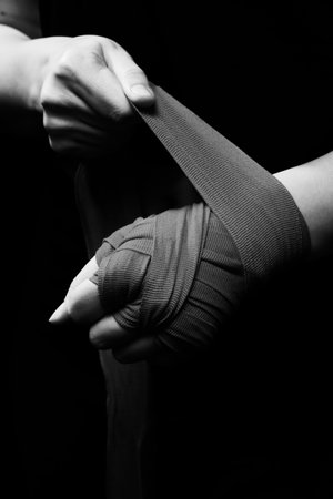 Muay Thai female boxer wearing strap on wrist closeup view. Fitness young woman preparing for boxing training on black background, monochromeの写真素材