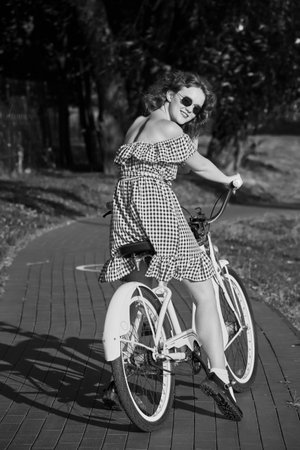 beautiful pinup girl with red dress and curly red hair moves on bike at sunny park in sun light, monochromeの写真素材