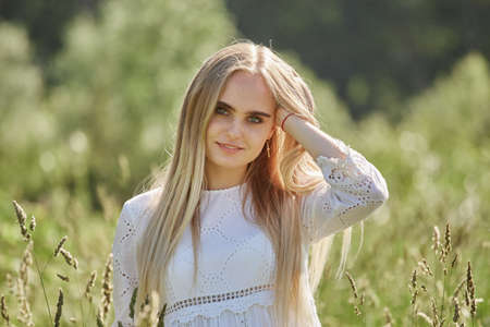 happy young blond woman in white dress at sunny meadow looking at camera smilingの写真素材