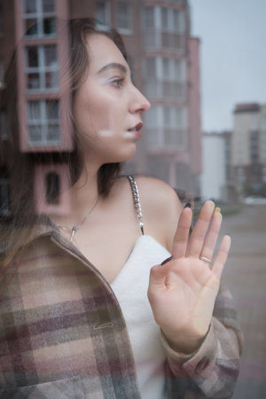 sad lonely young woman looking through window at modern cityの写真素材