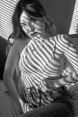 sensual young woman undressing in sun light with blinds shadows looking at camera, monochromeの写真素材