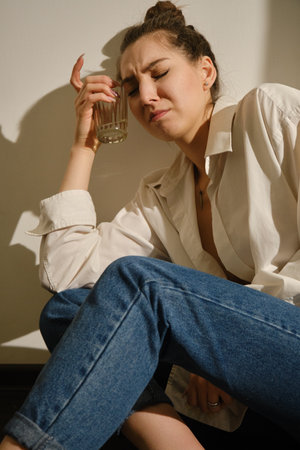 Young woman drinking alcohol from glass against sunny wall background, addiction