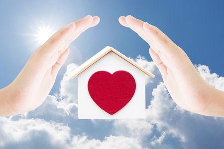 Two hands female protect house and red heart on blue sky clouds sun light background , home building landmark conceptの写真素材