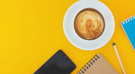 Top view. latte coffee in white cup with smartphone, notebook and pencil on yellow tableの写真素材