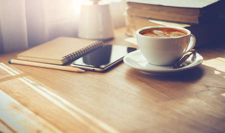 Closeup latte art coffee in white cup with smartphone and notebook on wooden table with sunlight through the window. Vintage color toneの写真素材
