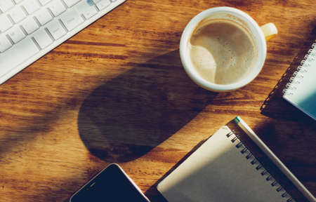 Top view. coffee cup, computer keyboard, mobile phone and notebook on wooden desk with sunlight. Vintage color toneの写真素材