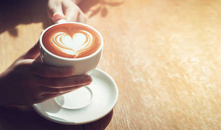 Close up girl hands holding latte art coffee in cup with white heart cream on top on wooden table. concept valentine coffeeの写真素材