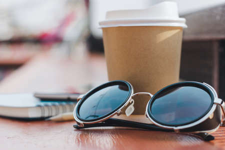 Close-up of retro sun glasses with paper cup of coffee and notebook, mobile phone on wooden counter, Concept relaxing after working outdoorの写真素材