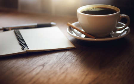 Closeup black coffee in cup with open blank notebook on wooden table. Vintage light and dark toneの写真素材