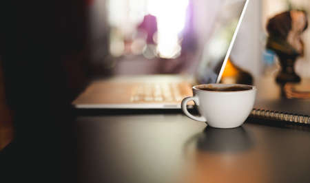 Close-up white coffee cup with notepad on desk and blurred laptop on background, Dark tone and color lightingの写真素材