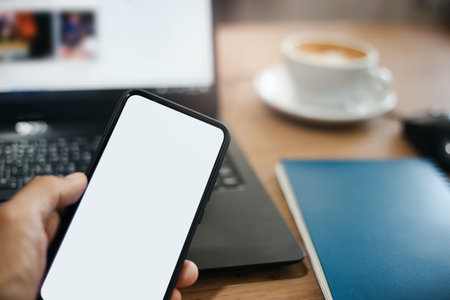 Selective focus on blank screen mobile in human hand with blur computer laptop and coffee cup on...