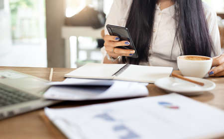 Close up. businesswoman using mobile phone and holding coffee cup while she working with laptop and paperwork of investmentの写真素材