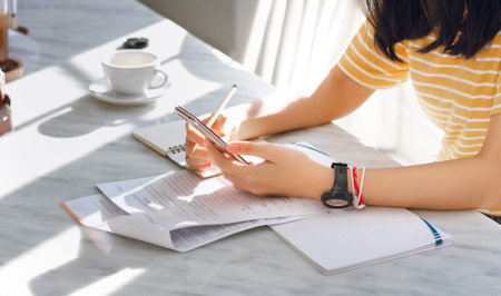 Young female using mobile phone to searching data online and working with paperwork on desk
