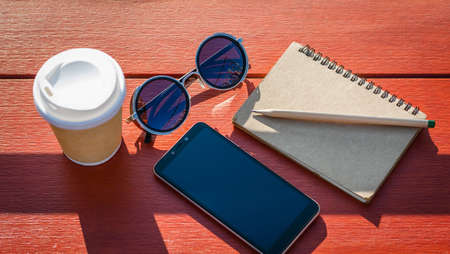 Top view. coffee cup, sunglasses, mobile phone and notebook on wooden desk with sunlight. concept people working outdoorsの写真素材
