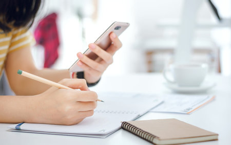 Closeup of hand, young female writing on notebook and using mobile phone to searching data online