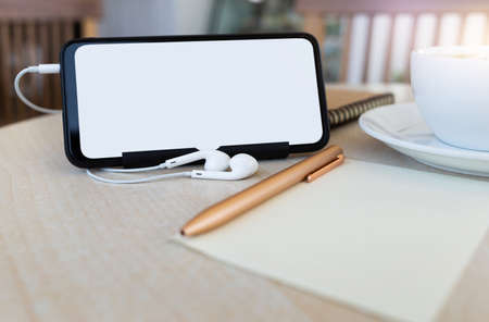 Business meeting Concept. blank screen smartphone with headphone, pen on note pad and cup of coffee on desk backgroundの写真素材