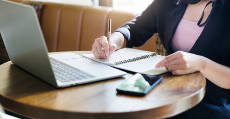 Focus on hand young asian businesswoman writing on book and working with computer laptop at home