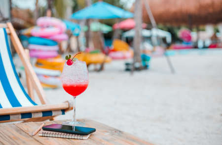 Close-up, juice with red cherry in champagne glass and mobile phone, book on wooden table on sand beach. Concept relaxing after work on summerの写真素材