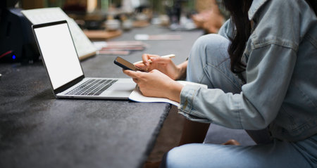 Focus on hand young asian woman holding mobile and using laptop working on counter in cafe. Concept...