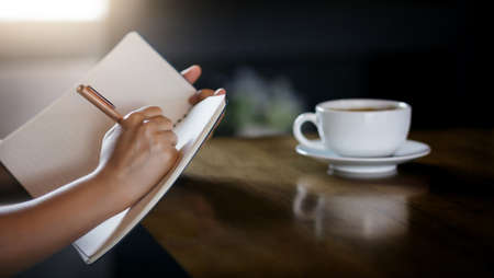 Close-up of young asian woman using pen writing on note book with white coffee cup on wooden table near window with sunlight shining through, Dark toneの写真素材