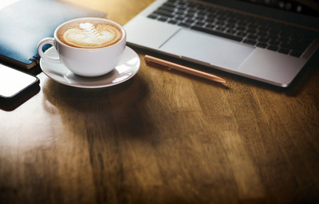 Focus on latte art coffee in white cup with computer laptop, mobile phone and blue note book on wooden tableの写真素材